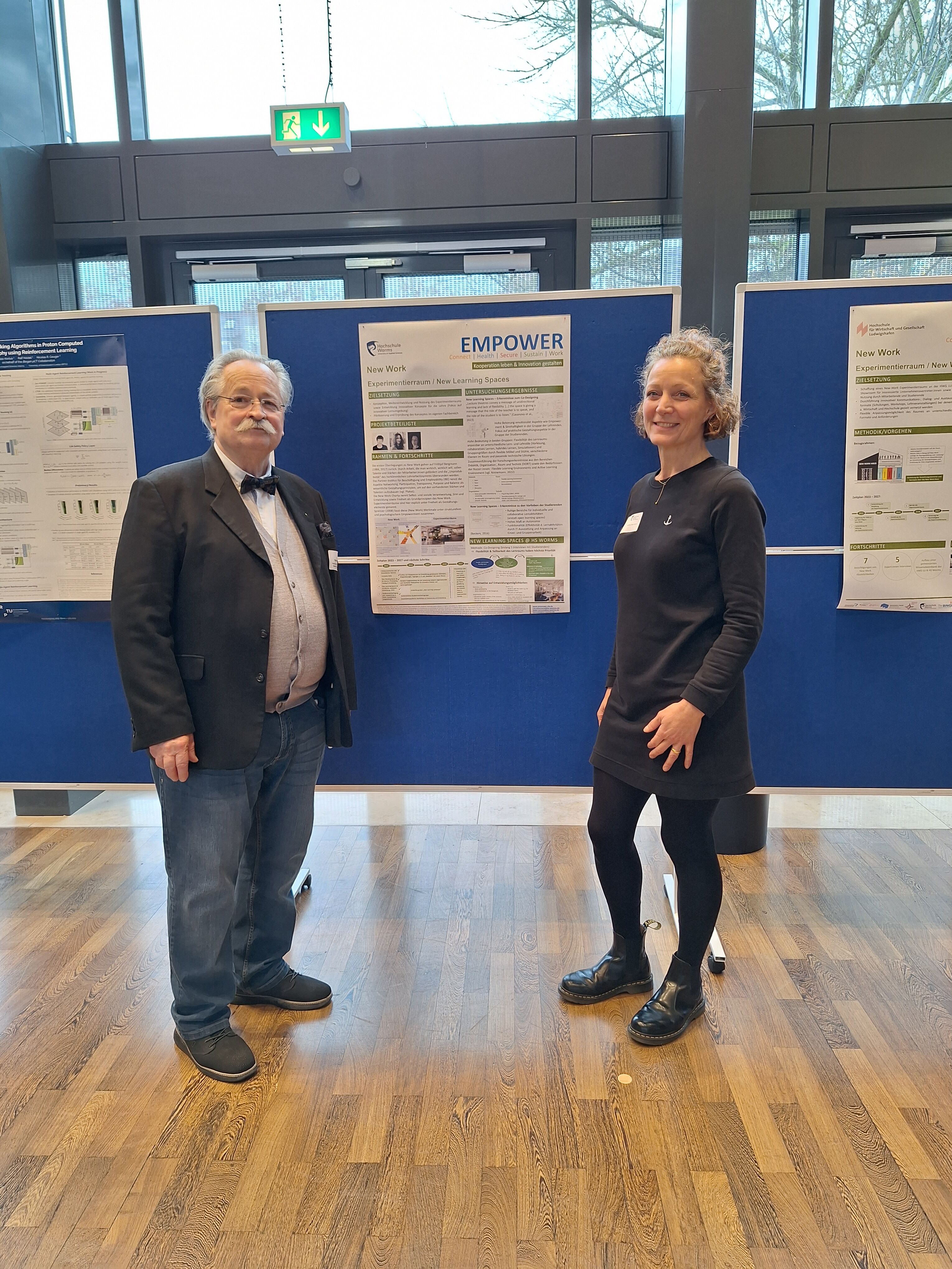 EMPOWER-Mitarbeiter Holger Roßberg und Sandra Debo-Scholz in der Aula der Hochschule Worms vor einem EMPOWER-Poster an einer blauen Pinnwand