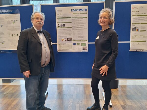 EMPOWER-Mitarbeiter Holger Roßberg und Sandra Debo-Scholz in der Aula der Hochschule Worms vor einem EMPOWER-Poster an einer blauen Pinnwand